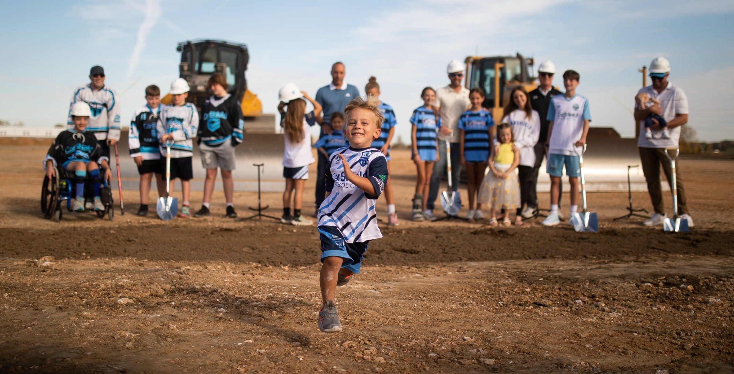 A young player in a CarShield jersey runs toward the camera at the CarShield Sportsplex groundbreaking, with teammates and hard-hat crews behind him